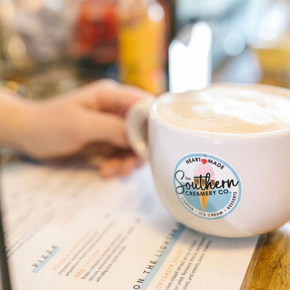 Latte in a branded Southern Creamery Co. cup sits on a café menu beside a blurred person at wooden counter.