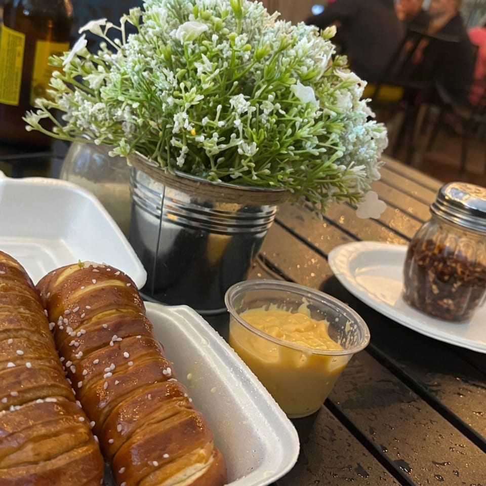 Two sesame-topped pretzel rolls sit in a white tray beside mustard, pickles, and a potted flower on a restaurant table.