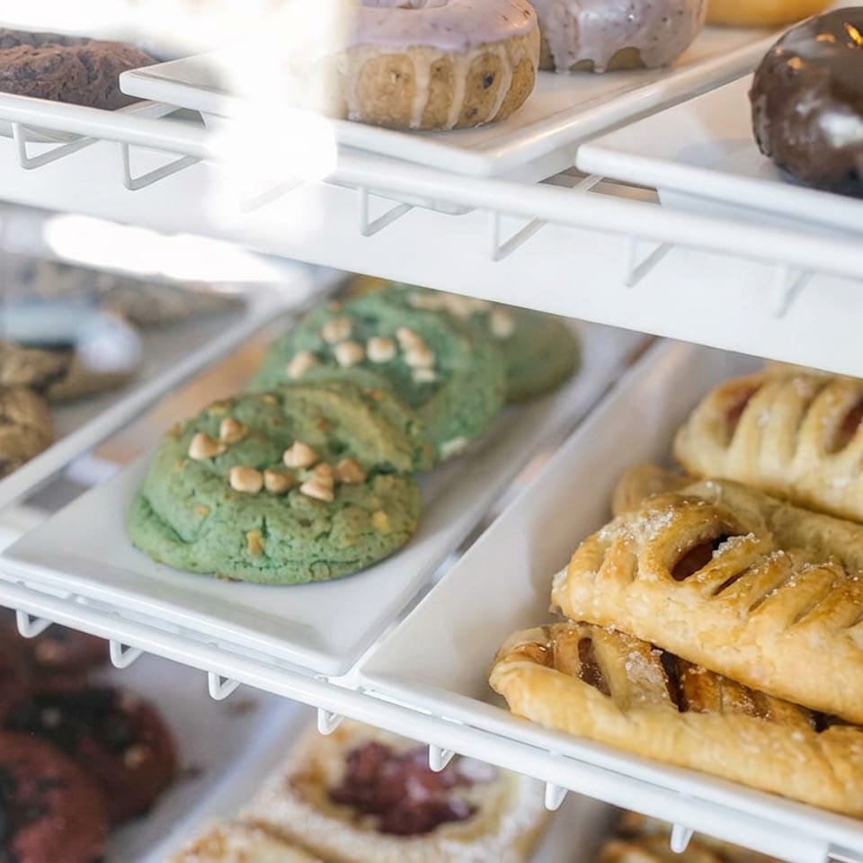 Pastries, cookies, colorful donuts, and fruit-filled hand pies arranged on white trays inside a bakery display case.