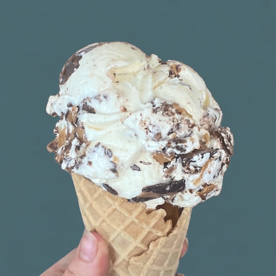 Hand holding a waffle cone piled with creamy cookies-and-cream ice cream against a teal background.
