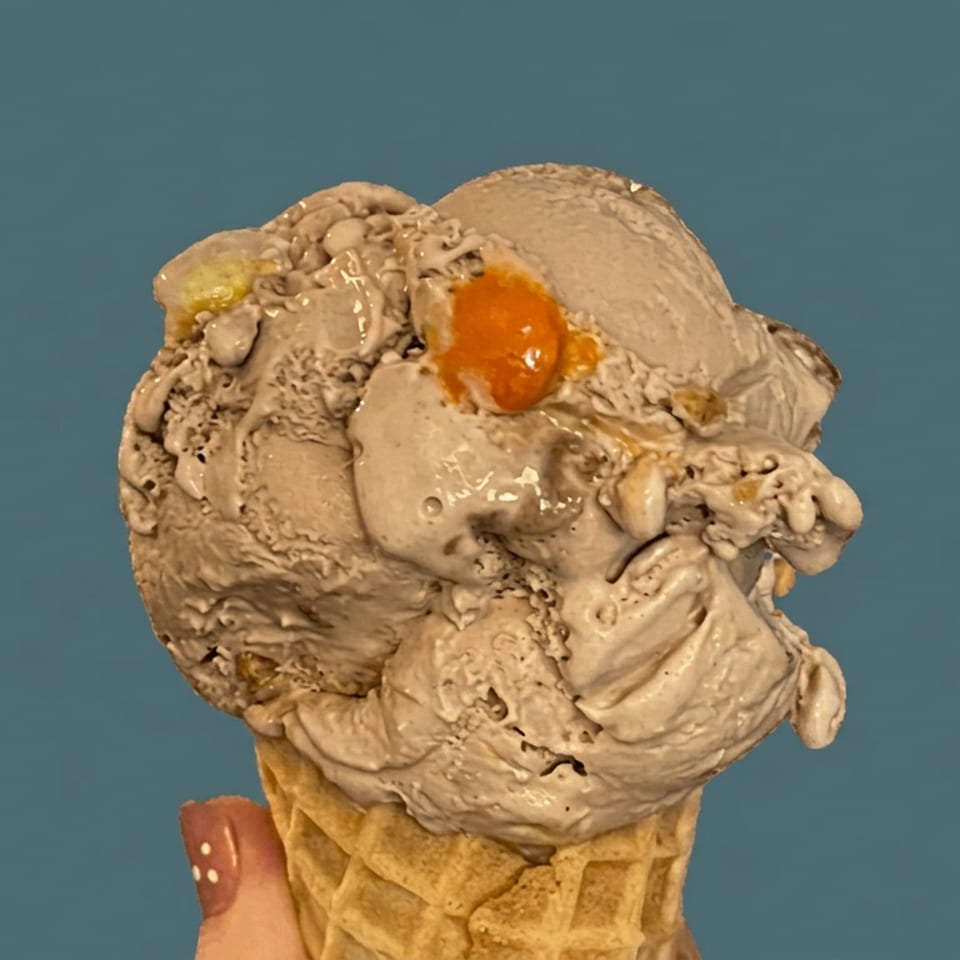 Waffle cone topped with an oversized scoop of melting beige ice cream and a bright orange garnish against a blue background.