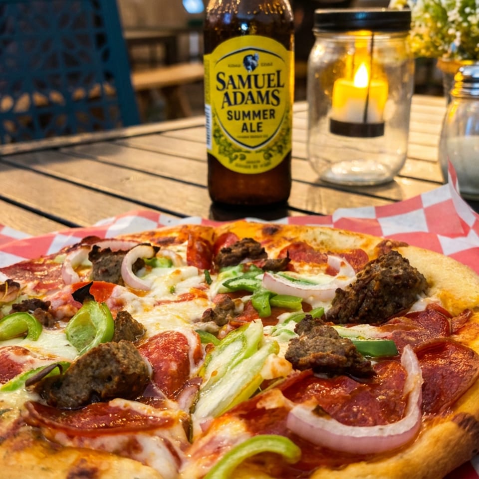 Loaded pepperoni pizza with peppers, onions, and sausage sits on a patio table beside a Samuel Adams Summer Ale and candle.