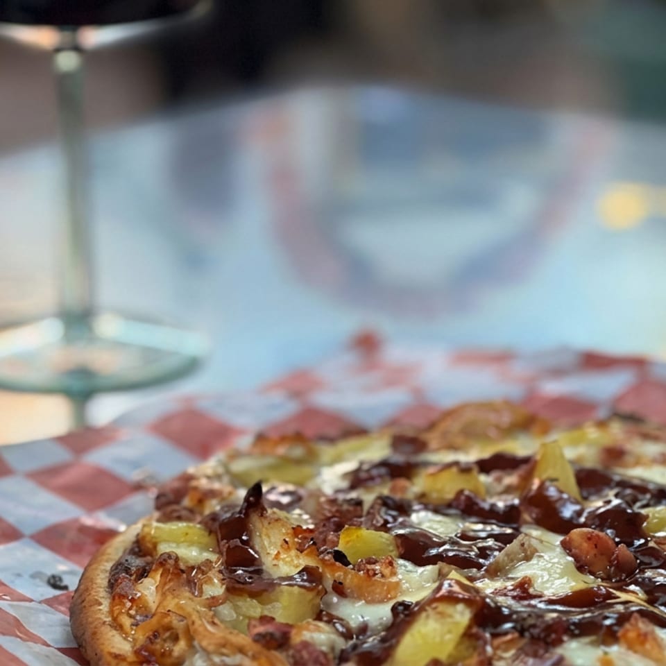 Loaded pizza with melted cheese, pineapple, mushrooms, and dark sauce on red-and-white checkered paper.